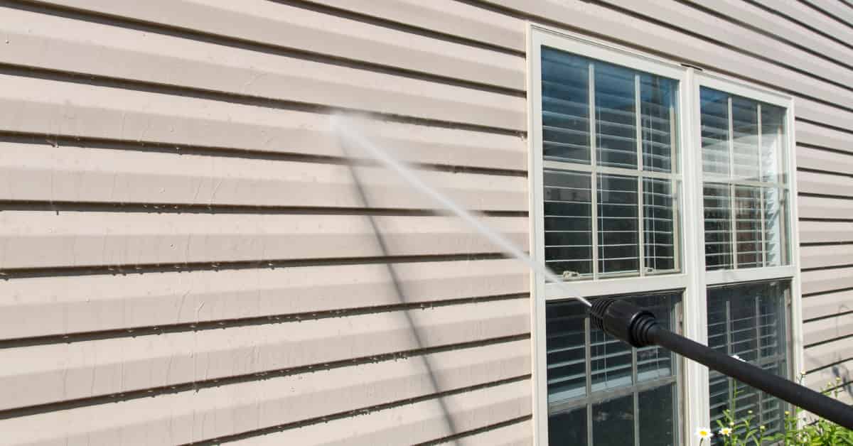 A high-pressure water cleaner is being used to wash the siding of a house, targeting a window.